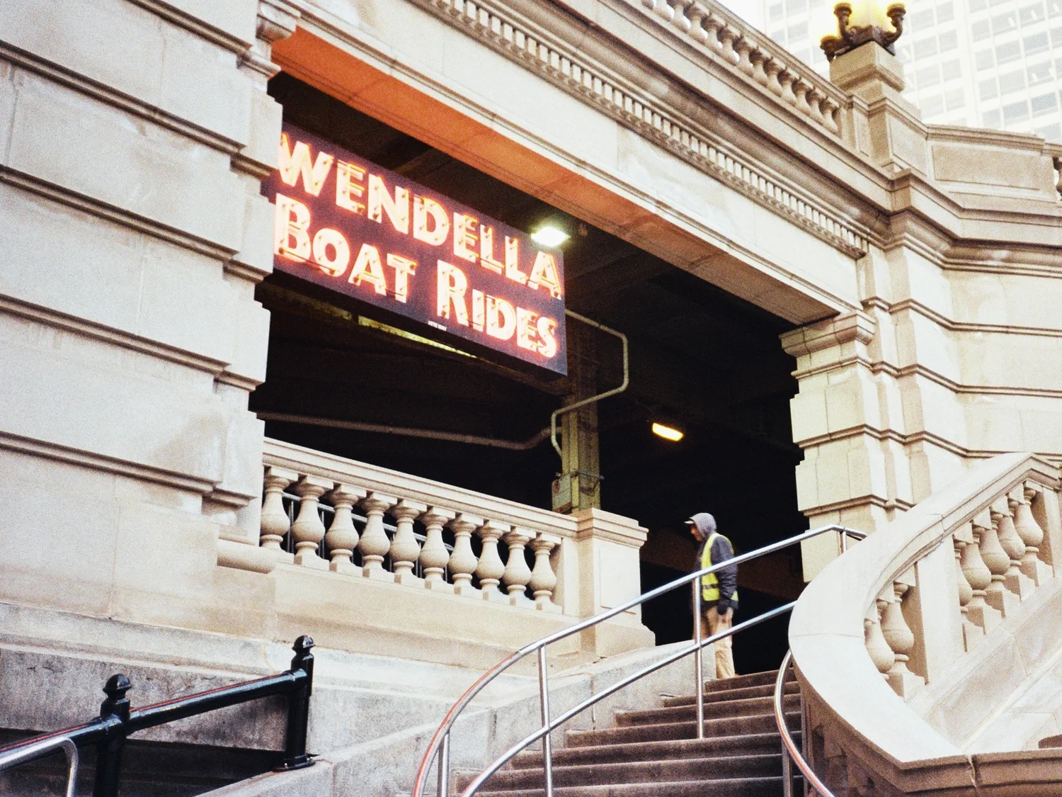A worker in a hi-vis vest climbs stone steps beneath the lit Wendella Boat Rides marquee on a Chicago bridgehouse