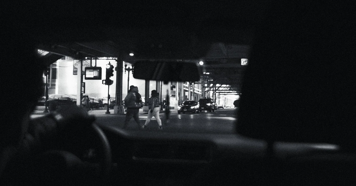 Black and white view from inside a car of two people crossing a city street at night beneath elevated train tracks