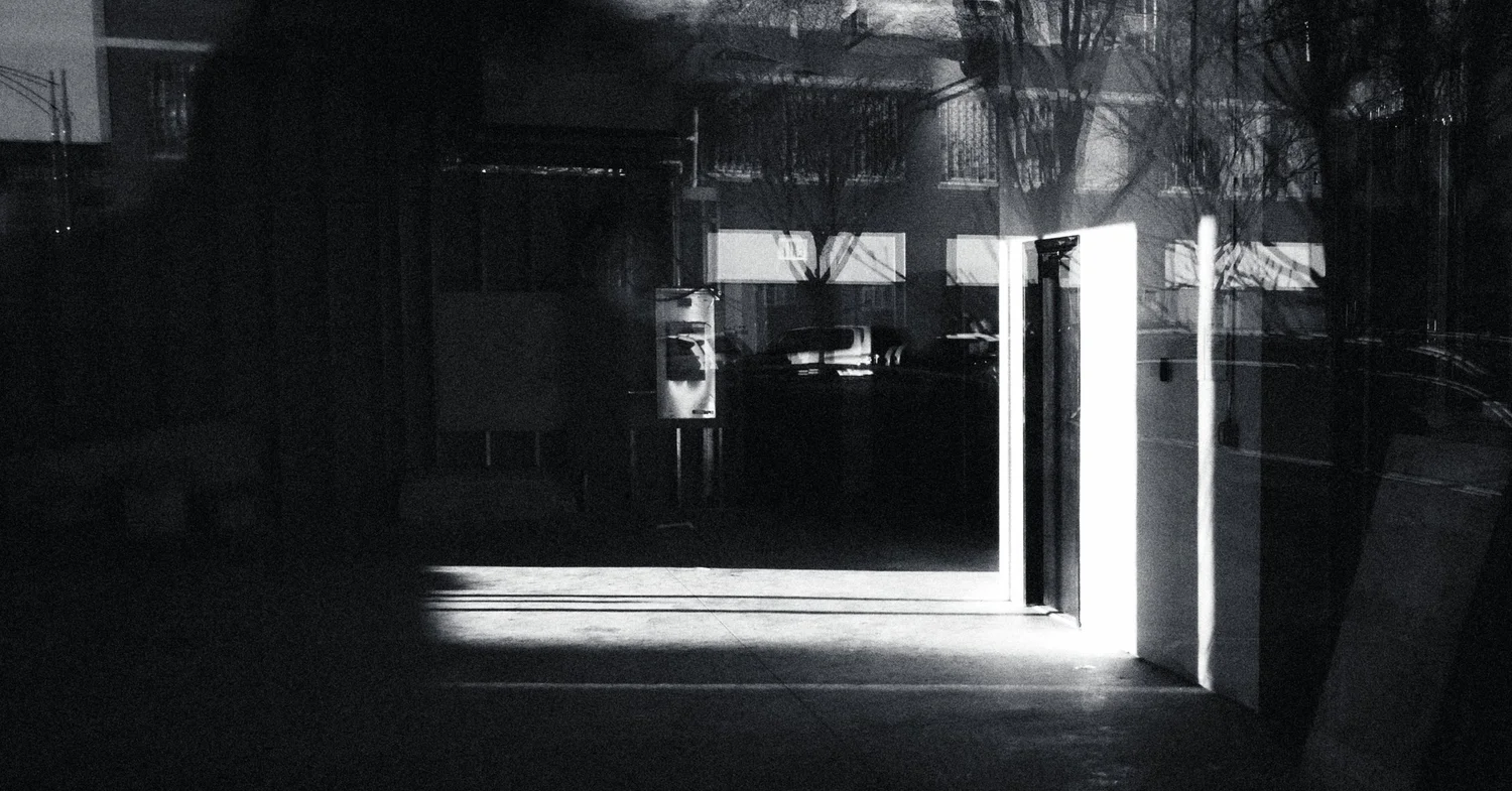 Black and white night scene of a brightly lit open doorway spilling light across a dark, empty street with bare trees reflected in glass