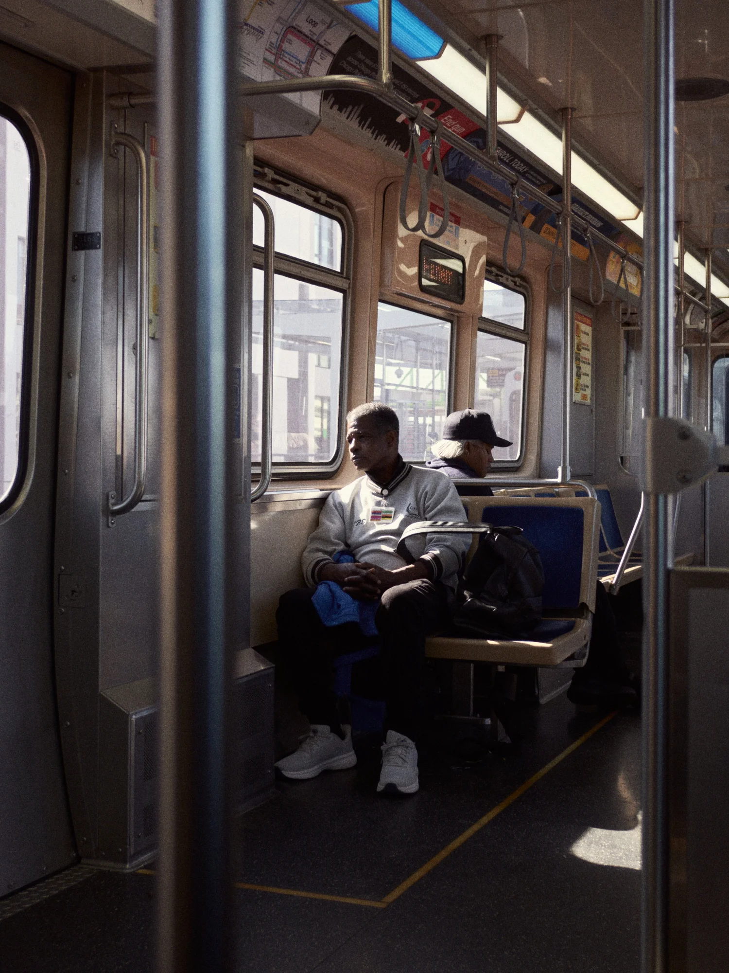 An older man sits in sunlight on a Chicago L train car, framed by chrome poles, with another passenger in a cap resting nearby