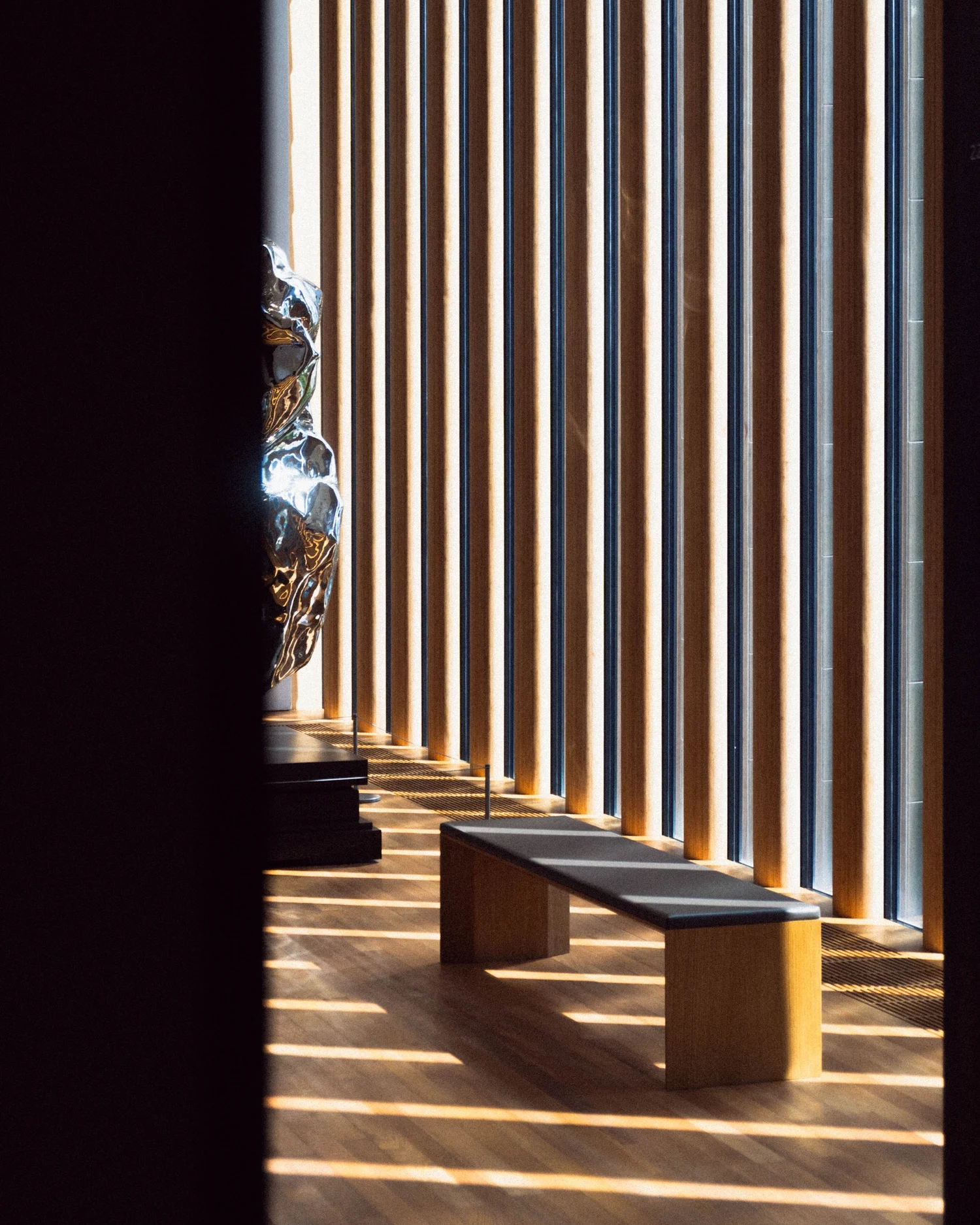 Late afternoon sun striping through tall wooden louvers onto a bench and a polished chrome sculpture in a quiet gallery
