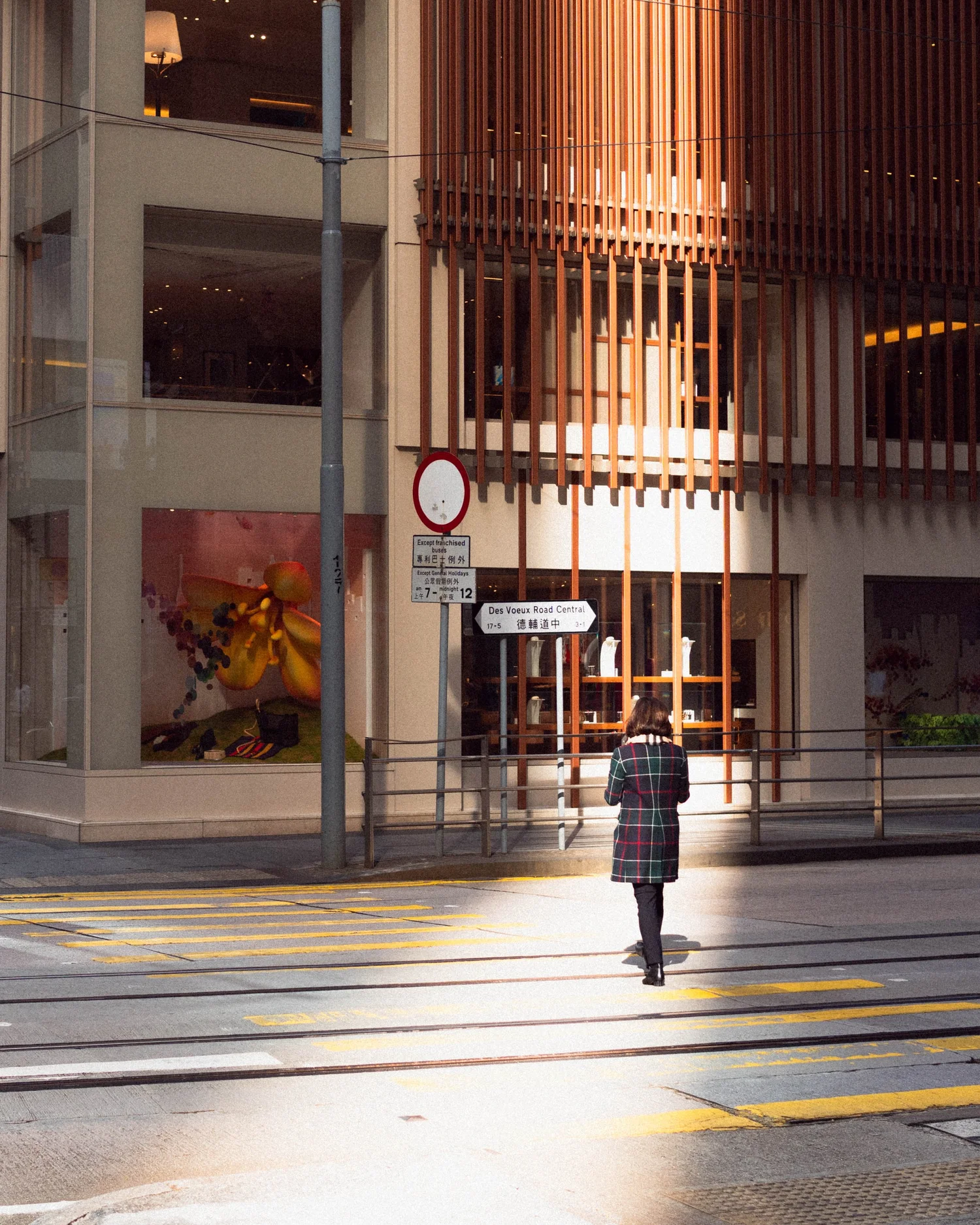 Street photograph of a person in a checked coat walking through a patch of warm sunlight in front of a wood slatted building in Hong Kong