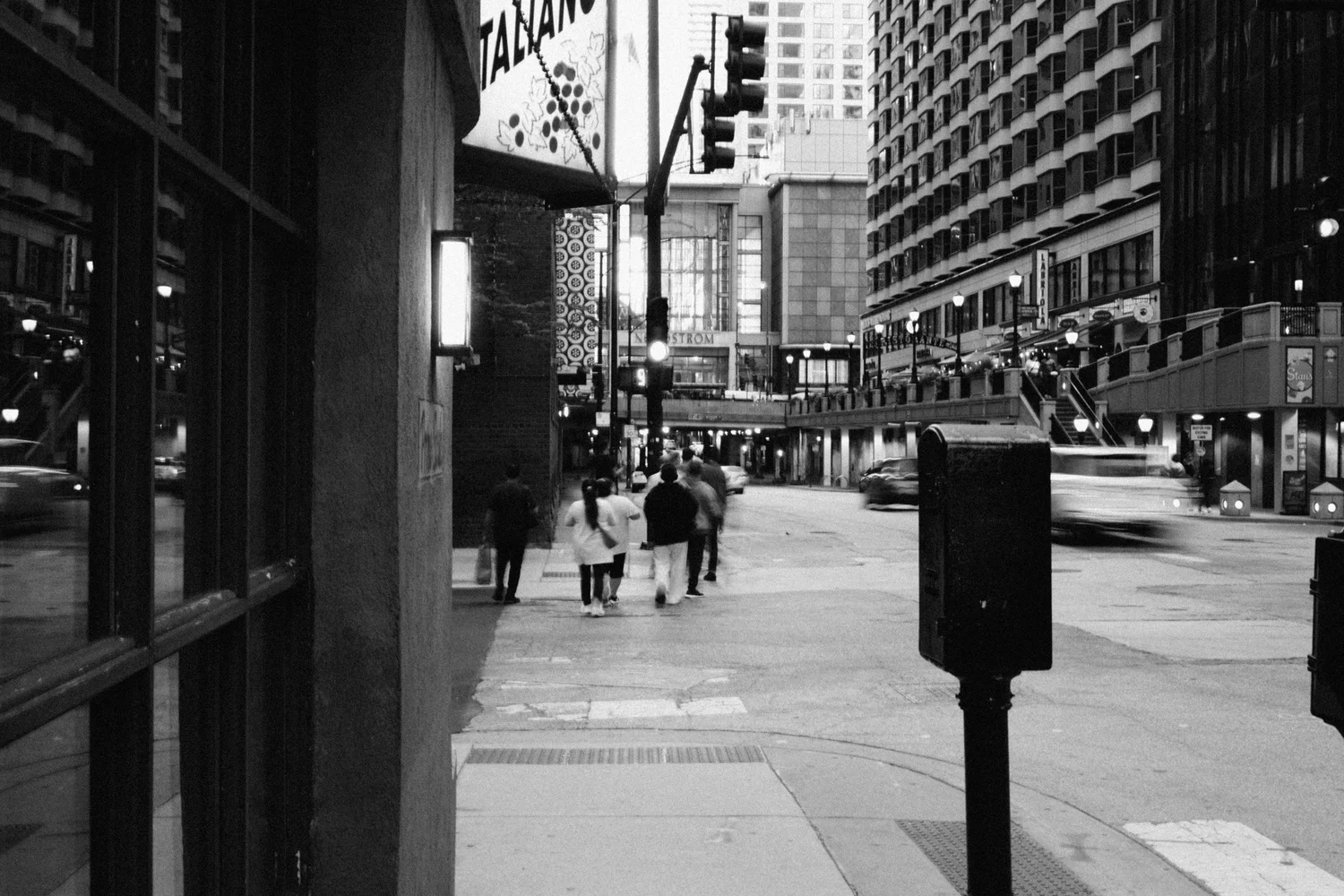 Black and white Chicago street at dusk with people walking away past an Italian restaurant sign and a motion-blurred passing car
