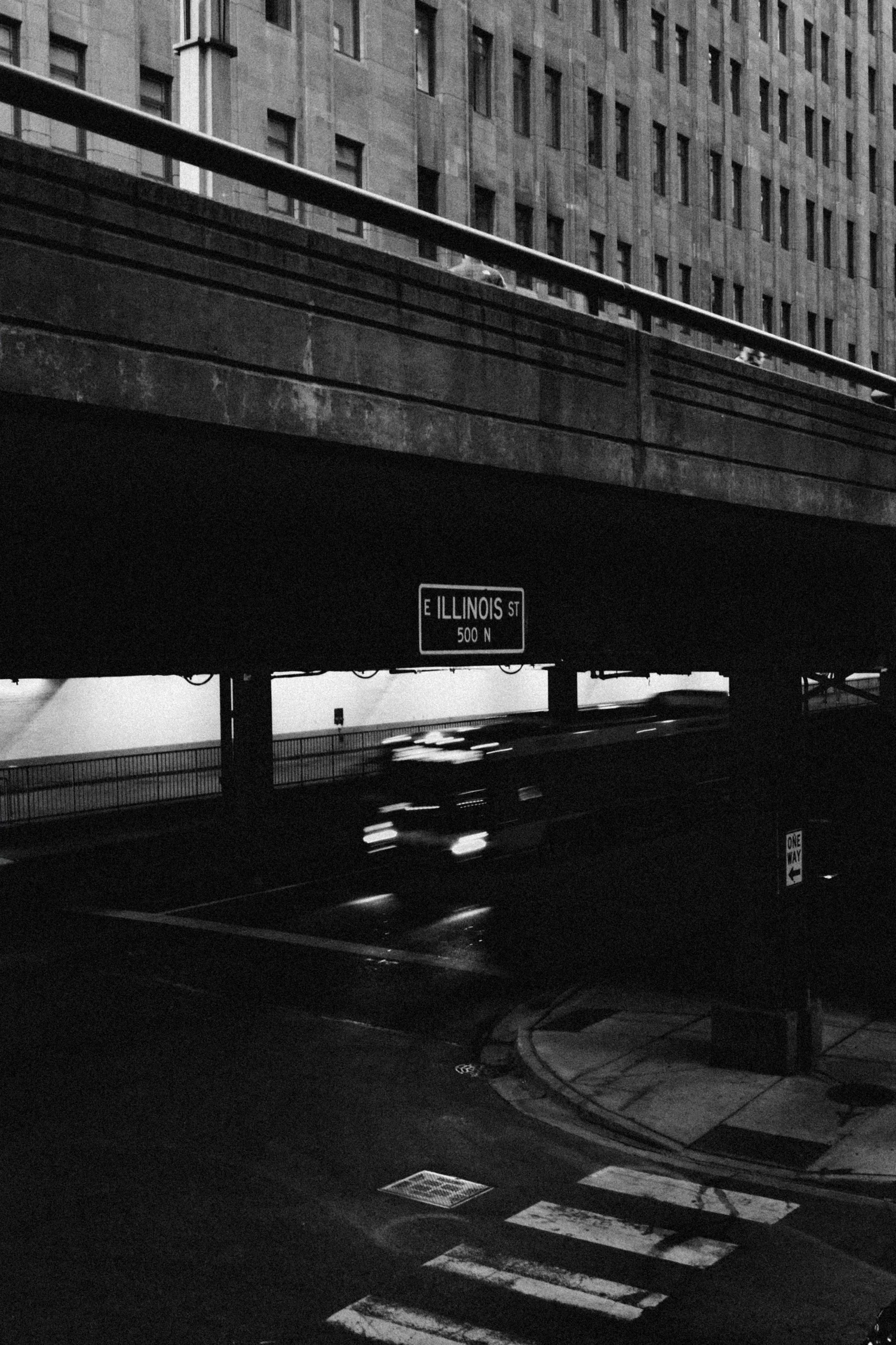 Black and white view under a Chicago bridge overpass, an E Illinois St sign and a motion-blurred car passing toward the lakefront