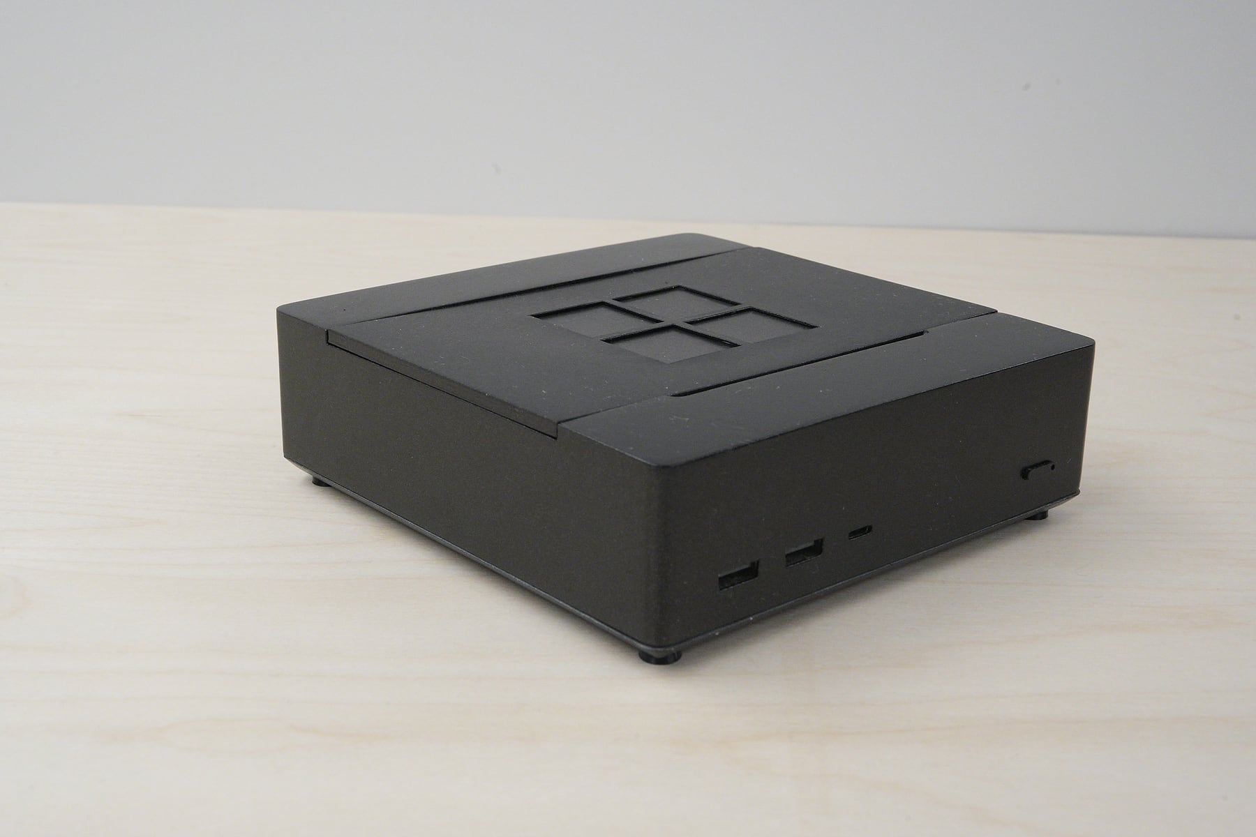 The physical prototype on a light wood desk, seen from a front corner. It is a low black box with a four pane logo on its lid, two USB A ports and a USB C port on the front and a small power button at the right.