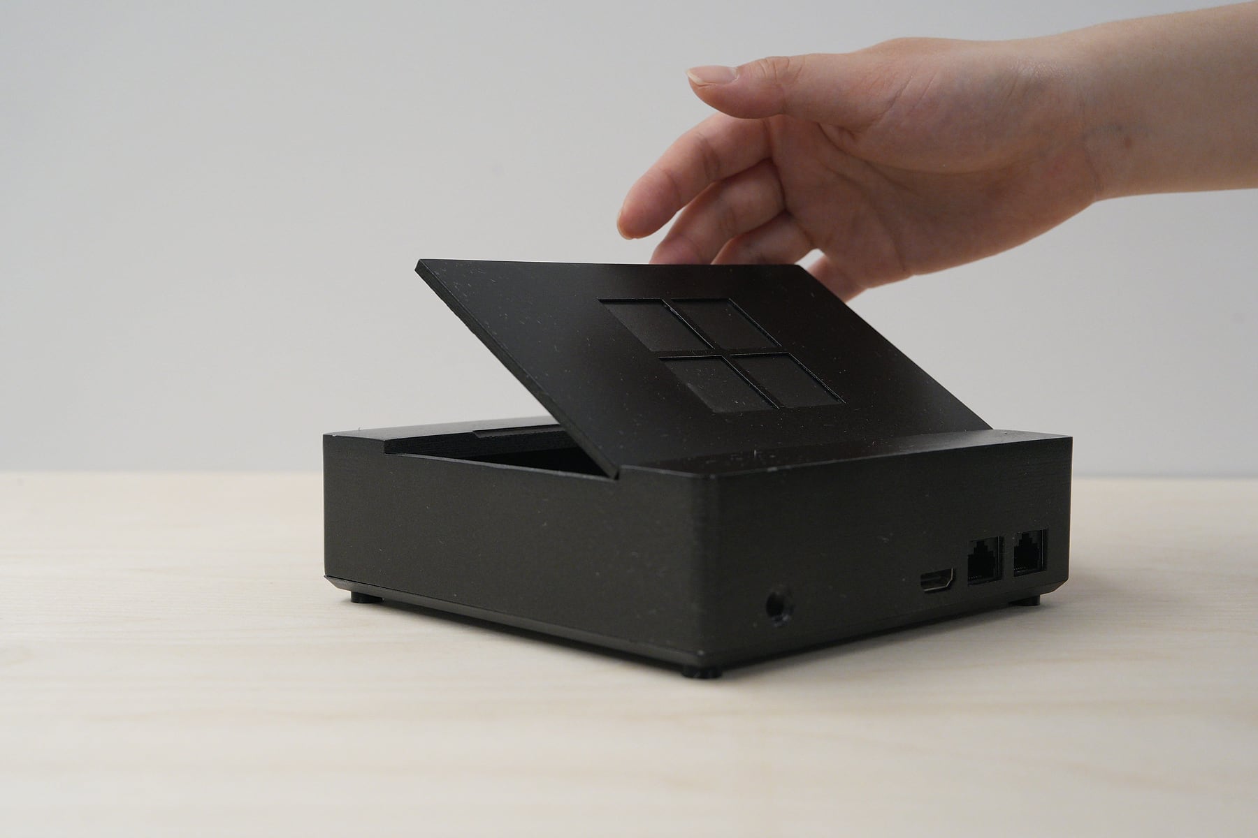 A hand lifting the lid of the prototype, seen from the back. The lid tilts up to reveal the empty drive bay, and the back of the unit shows a power jack, an HDMI port and two ethernet ports.
