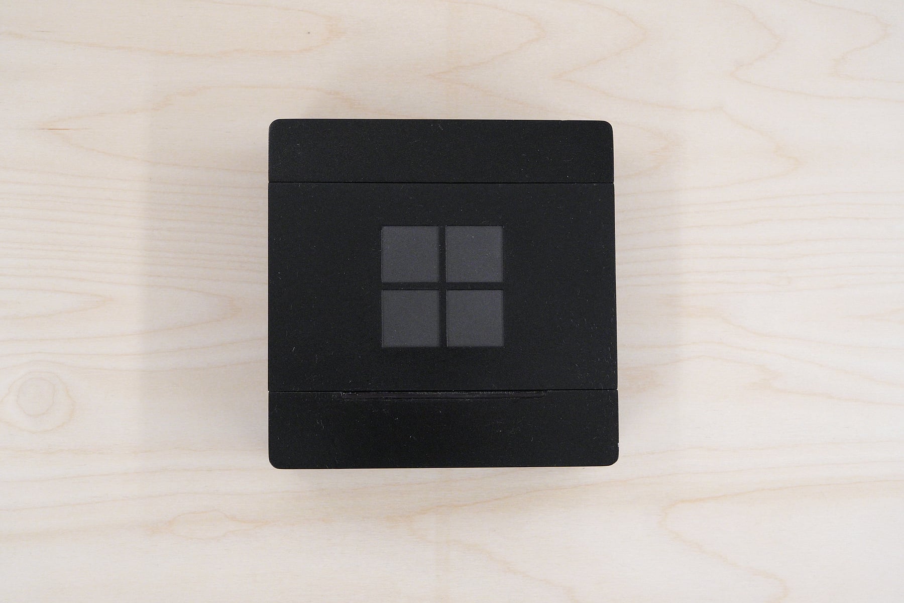 The prototype seen from directly above on a light wood surface. A black square lid carries the four pane logo in slightly lighter grey, with a narrow seam at each end.
